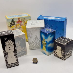 May include: A collection of vintage perfume boxes in various shapes and sizes. The boxes feature illustrations of figures, floral patterns, and text. Colors include blue, white, black, and yellow. A coin is in the foreground.