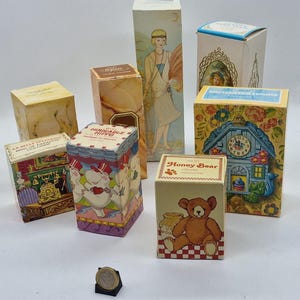 Peut inclure: Une collection de flacons de parfum vintage avec différents motifs. Les flacons sont dans des boîtes avec des illustrations colorées d'animaux, de fleurs et d'une tour d'horloge. Les boîtes sont sur une surface blanche.