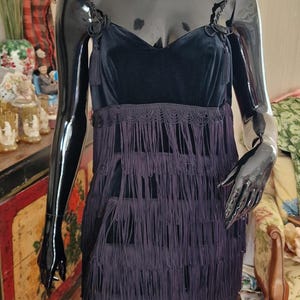 Peut inclure: Une robe en velours noir avec une frange sur le bas. La robe a un décolleté en cœur et des bretelles fines. La frange est superposée et descend jusqu'aux genoux.