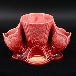 May include: A pink ceramic vase with three openings, each with a scalloped rim. The vase has a textured surface and a decorative design.