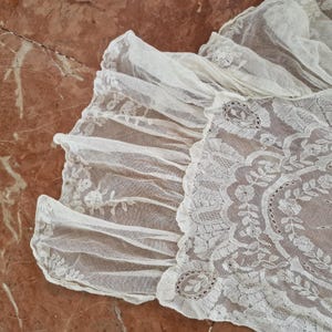 May include: Close-up of an off-white lace textile with detailed floral and geometric patterns. The lace has a ruffled edge, suggesting a vintage or antique piece. The fabric has a soft, airy texture. The background is a mottled, reddish-brown surface.