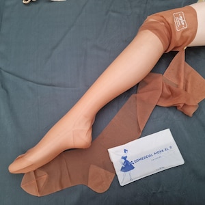 Puede incluir: Medias de nailon color piel, de aspecto vintage, parcialmente puestas. Se aprecia la transparencia del tejido. Un paquete blanco con el texto "COMERCIAL MOYA EL 9" y un dibujo de una mujer es visible.