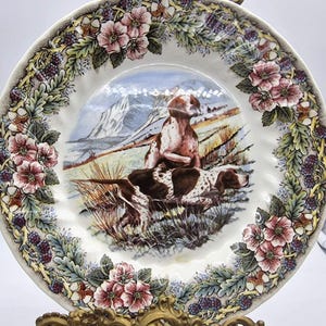 Könnte beinhalten: Dekorative Platte mit einer Jagdszene mit zwei Hunden und einer Person auf einem Feld mit Bergen im Hintergrund. Die Platte hat einen floralen Rand mit rosa Blüten, grünen Blättern und lila Beeren.