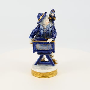 Op de afbeelding: Een porseleinen beeldje van een jongen in een blauwe jas en hoed die een handorgel bespeelt. Een kleine aap zit op zijn schouder. Het beeldje is versierd met gouden accenten.