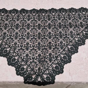 Könnte beinhalten: Ein schwarzer Spitzenschal mit gewelltem Rand. Der Schal weist ein sich wiederholendes Blumenmuster in seinem gesamten Design auf. Die Spitze ist von dunkler, einfarbiger schwarzer Farbe, und der Schal liegt auf einer hellrosa, floral gemusterten Oberfläche.