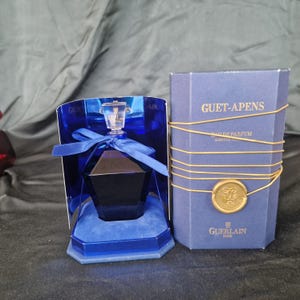 Puede incluir: Un frasco de perfume azul oscuro con un diseño facetado, exhibido en una caja forrada de terciopelo azul. La caja está junto a una caja azul a juego con el texto "GUET-APENS" y "GUERLAIN PARIS". La caja está sellada con un emblema dorado y atada con hilo dorado.