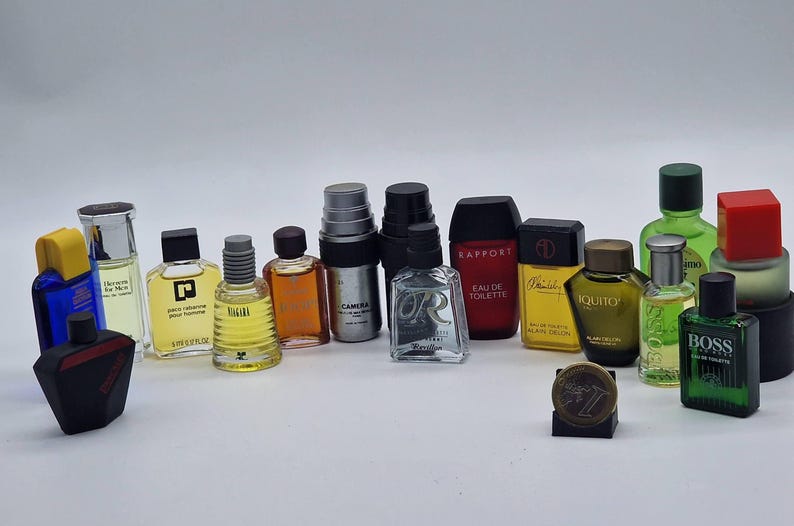 May include: A collection of miniature perfume bottles in various shapes, sizes, and colors. The bottles have different colored caps and labels, with some featuring text. The bottles are arranged on a white surface.