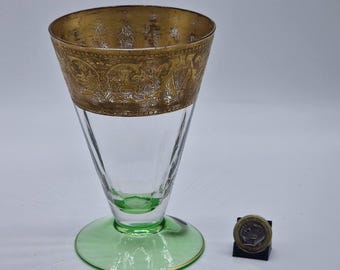May include: A clear glass goblet with a gold-colored rim featuring an ornate design. The goblet has a green base and a conical shape. A coin is placed next to the goblet for scale.
