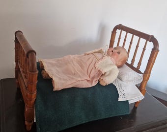 Könnte beinhalten: Eine Puppe mit einem rosa Kleid und einem weißen Pullover liegt auf einem Holzbett mit einer grünen Decke und einem weißen Kissen. Das Bett hat einen Holzrahmen mit einem Lattenrost.