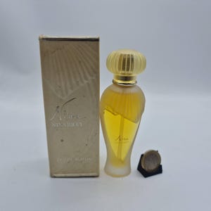 Könnte beinhalten: Eine Flasche Nina Ricci Eau de Parfum mit goldfarbenem Verschluss und passender Schachtel. Die Flasche ist mit einer gelben Flüssigkeit gefüllt und trägt den Namen "Nina". Die Schachtel ist beige und trägt den Namen "Nina Ricci".