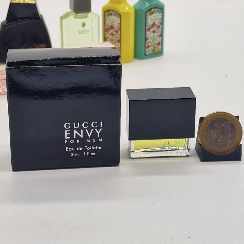 May include: A miniature perfume bottle and box set. The black box reads "GUCCI ENVY FOR MEN Eau de Toilette 3 ml .1 fl.oz." The perfume bottle is a small, clear glass cube with a black cap. A coin is next to the perfume.
