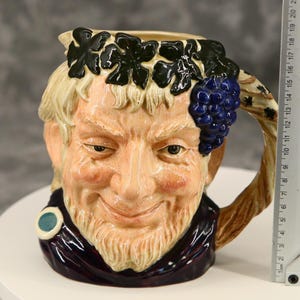 May include: A ceramic mug depicting a smiling, bearded man adorned with a grape cluster and leaf crown. The mug, with a handle, is painted in tan, brown, and purple hues. The mug is approximately 18 cm tall.