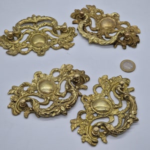 Könnte beinhalten: Vier verzierte, goldfarbene Metall-Schubladengriffe mit aufwendigen floralen Designs. Jeder Griff hat eine kreisförmige Mitte und einen dekorativen Schleifen-Griff.