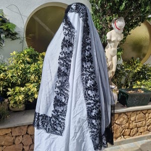 May include: A black lace veil with a floral pattern. The veil is draped over a white fabric and is partially covering a statue.