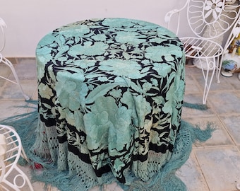 Peut inclure: Un châle noir orné de broderies florales vert clair sur toute la surface. Le châle présente de longues franges vert clair sur les bords et est drapé sur une table ronde, s'étalant sur le sol carrelé en dessous.
