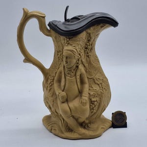 May include: A tan ceramic jug with a dark grey lid and handle. The jug has a relief carving of a seated figure and grape vines. The jug is likely vintage and decorative, with a height of approximately 20 cm.