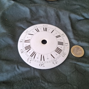 Può includere: Un quadrante bianco con numeri romani neri e indicatori dei minuti. Il quadrante ha un foro centrale ed è accanto a una moneta da un euro. Il quadrante è probabilmente un pezzo di ricambio o per un progetto artigianale.