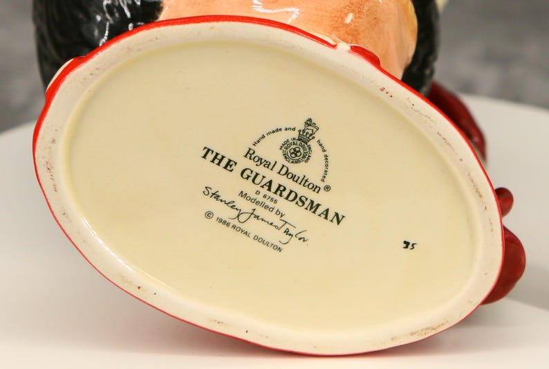 Może przedstawiać: Podstawa ceramicznej figurki w kolorze kremowym z czerwonym obramowaniem, z czarnym napisem "THE GUARDSMAN" i "Royal Doulton". Model autorstwa Stanley James Taylor.