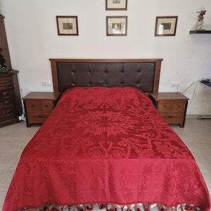 May include: A bedroom set with a dark brown headboard and matching bedside tables. The bed is covered with a vibrant red bedspread with a detailed floral pattern and fringe trim. The headboard has a tufted leather center, creating a focal point.