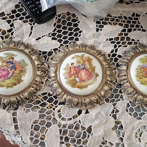 Pode incluir: Três molduras ornamentadas em tom dourado com desenhos intrincados, cada uma com uma placa redonda de porcelana branca com uma cena pintada de um casal em um ambiente de jardim.