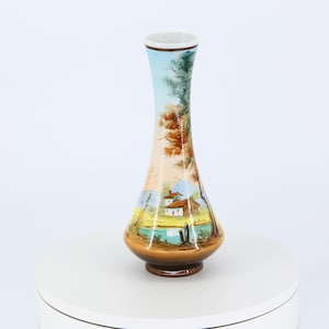 以下が含まれることがあります： 湖畔の家と木々が描かれた茶色の陶器の花瓶。花瓶は首が細く、底が広い。