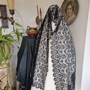 May include: A black lace mantilla, a traditional Spanish head covering, draped over a white form. The intricate floral pattern of the lace is visible against the white background. The mantilla is a formal accessory often worn for religious or cultural events.