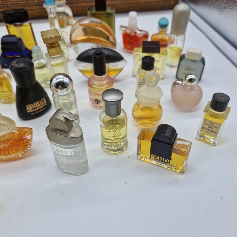 K&ouml;nnte beinhalten: Eine Sammlung von Miniatur-Parf&uuml;mflaschen in verschiedenen Formen und Farben. Die Flaschen haben unterschiedliche Verschlussdesigns und enthalten Fl&uuml;ssigkeiten in Gelb-, Orange- und Klart&ouml;nen. Einige Flaschen haben Etiketten, angeordnet auf einer wei&szlig;en Oberfl&auml;che.