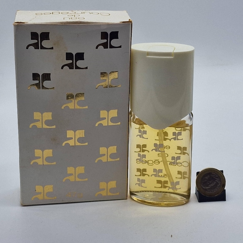 May include: A vintage Courr&egrave;ges Eau de Parfum bottle with a white cap and a matching box. The box and bottle are adorned with a repeating gold logo pattern. The perfume bottle contains a light amber liquid.
