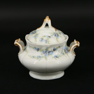 Pode incluir: Açucareiro de porcelana branca com tampa, adornado com padrões florais azuis e detalhes dourados. O açucareiro tem duas pegas e um adorno decorativo na tampa. Uma peça clássica para servir açúcar.
