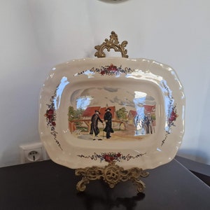 Puede incluir: Plato rectangular de cerámica color crema con una escena pintada a mano de personas con abrigos y sombreros negros. El plato tiene un borde festoneado con detalles florales y de vid. Se exhibe sobre un soporte decorativo de latón.