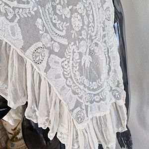 May include: Cream-coloured lace shawl with intricate floral and leaf patterns. The delicate fabric features a scalloped edge and a ruffled trim. The shawl is draped over a black mannequin, showcasing its elegant design.