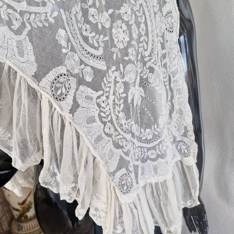 May include: Cream-coloured lace shawl with intricate floral and leaf patterns. The delicate fabric features a scalloped edge and a ruffled trim. The shawl is draped over a black mannequin, showcasing its elegant design.