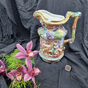 Puede incluir: Una jarra de cerámica colorida con un diseño en relieve detallado que presenta figuras, uvas y follaje. El asa y el borde están adornados con racimos de uvas y hojas. La jarra tiene una base azul claro y un interior blanco.