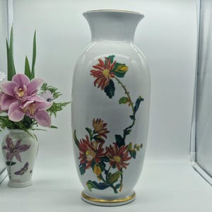 Op de afbeelding: Een hoge, witte keramische vaas met een gouden rand, versierd met rode en gele chrysantenbloemen en groene bladeren. Links staat een kleinere vaas met roze bloemen. De vaas is ongeveer 30 cm hoog.