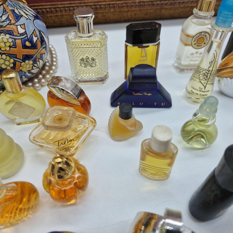 K&ouml;nnte beinhalten: Eine Sammlung von Miniatur-Parf&uuml;mflaschen in verschiedenen Formen und Gr&ouml;&szlig;en. Die Flaschen sind mit unterschiedlich farbigen Fl&uuml;ssigkeiten gef&uuml;llt, darunter Gelb, Orange und Klar. Einige Flaschen haben aufwendige Designs und Etiketten, andere sind schlicht und elegant.