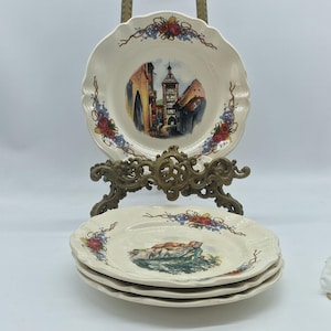 Puede incluir: Un juego de platos decorativos de color crema con bordes festoneados. Cada plato presenta una escena de paisaje central y detalles florales y de vid. Un plato se exhibe en un soporte decorativo de latón, con tres platos adicionales apilados debajo.