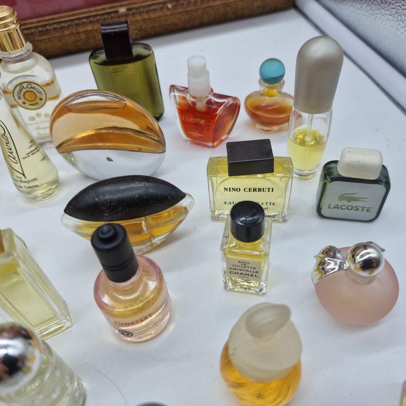 K&ouml;nnte beinhalten: Eine Sammlung von Miniatur-Parf&uuml;mflaschen in verschiedenen Formen und Farben. Die Flaschen bestehen aus klarem, bernsteinfarbenem und farbigem Glas mit verschiedenen Verschlussdesigns. Einige Flaschen zeigen Markennamen wie Chanel und Lacoste.