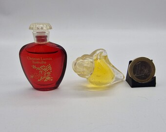Puede incluir: Una botella de perfume roja Christian Lacroix Tumulte con tapón transparente, una botella de perfume de vidrio amarillo con forma de pájaro y una moneda de un euro sobre un soporte negro. La botella de perfume tiene detalles florales dorados.