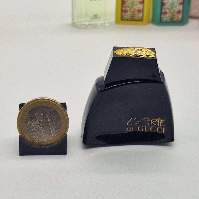 May include: A black perfume bottle with gold accents and the text "L'Arte di Gucci" in gold lettering. A one euro coin is next to the bottle in a black holder. Other perfume bottles are in the background.