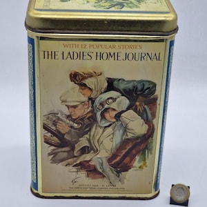 Könnte beinhalten: Eine rechteckige Vintage-Blechdose mit goldenem Deckel und blauen Seiten. Die Vorderseite zeigt eine Illustration aus The Ladies' Home Journal, August 1914. Das Bild zeigt Personen in Vintage-Kleidung und -Brillen. Die Dose hat einen goldenen Rand.