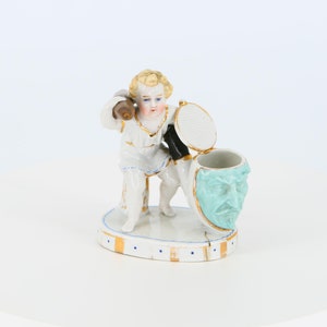 May include: A porcelain figurine depicting a cherubic figure holding a mallet, with a hinged container featuring a teal face design. The figurine is white with gold and blue accents, set on a white base with a gold and blue border.