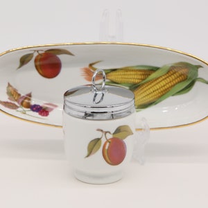 May include: A white porcelain egg coddler with a silver lid and a single red plum design. The coddler is sitting on a white oval serving dish with a gold rim and a design of corn on the cob and a red plum.