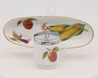 Könnte beinhalten: Ein weißer Porzellan-Eierwärmer mit einem silbernen Deckel und einem roten Pflaumen-Design. Der Eierwärmer steht auf einer weißen ovalen Servierplatte mit goldfarbenem Rand und einem Design aus Maiskolben und einer roten Pflaume.