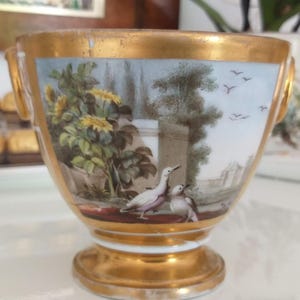 Pode incluir: Uma chávena de porcelana com rebordo, base e pegas laterais pintadas a ouro. O exterior apresenta uma cena pintada à mão de dois pássaros junto a um muro de pedra, rodeados por folhagem verde, flores amarelas e um céu azul claro com pássaros.