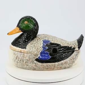 Puede incluir: Un plato de servir de cerámica con forma de pato y tapa. El pato está pintado en tonos marrones, negros, verdes y azules. El plato es perfecto para servir una variedad de alimentos.