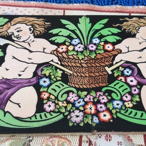 Peut inclure: Deux chérubins, l'un tenant une faux et l'autre une pelle, sont représentés sur un carreau coloré avec un panier de fleurs au centre. Le carreau présente un fond noir avec un feuillage vert et des accents floraux.