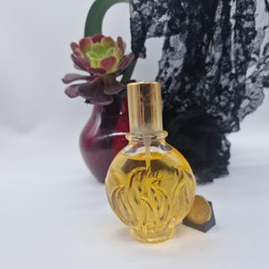 Könnte beinhalten: Ein runder, klarer Glasparfümflakon mit goldener Flüssigkeit, der an der Basis ein strukturiertes, flammenartiges Design und einen goldenen Verschluss aufweist. Auf der Vorderseite steht das Wort "Fidji". Dahinter sind eine Sukkulente und schwarze Spitze.