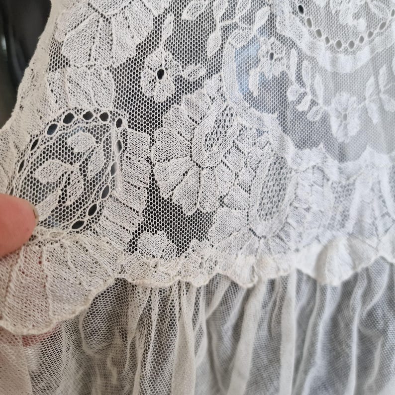 May include: Close-up of delicate cream-coloured lace fabric. The lace features floral and leaf patterns, with a scalloped edge and a ruffled trim. The intricate design is visible against a dark background, highlighting the texture and detail of the textile.