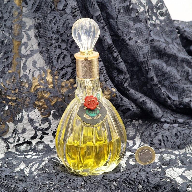 May include: A vintage perfume bottle with a clear, ribbed glass body and a decorative, clear stopper. The bottle has a gold-colored neck and a red seal with the number "4711". The perfume inside is yellow. A coin is next to the bottle.
