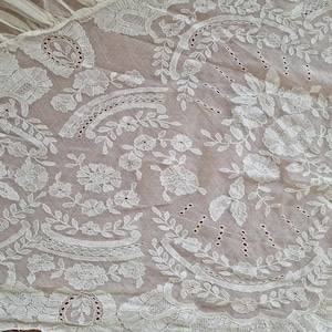 May include: Detailed view of white lace fabric with floral and leaf motifs. The lace showcases elaborate embroidery and openwork, giving a textured and airy feel. The fabric is likely used for decorative purposes, such as clothing or home decor.